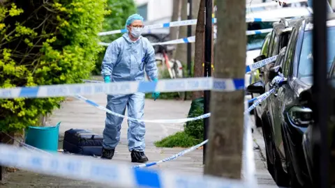 Police tape crosses a street between trees as a forensics worker walks down it