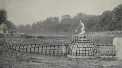 The Builder's Journal A historic black-and-white photograph showing the underwater room during construction. The glass-domed structure and connecting tunnel are visible before the lake was filled with water. A statue stands on top of the dome and open ground surrounds the site.