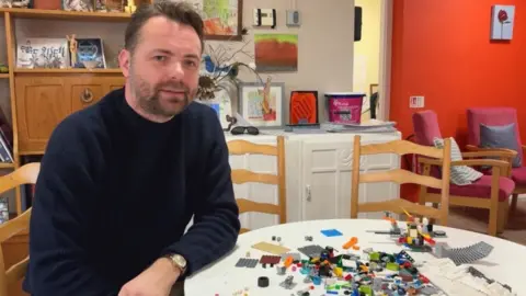 A man sits at a table full of Lego. He is wearing a navy blue jumper and has short brown hair. There is a sideboard and some chairs in the background. The Lego on the table is a collection of individual, multi-coloured bricks.