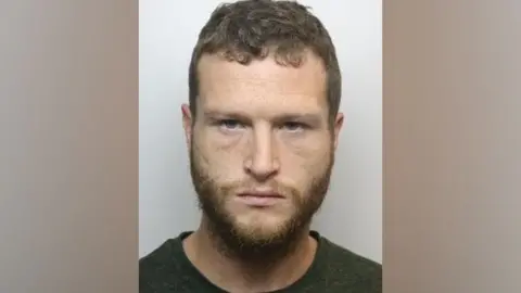 South Yorkshire Police Levi Fishlock is shown in a police mug shot. With short brown hair and beard he is looking straight at the camera. He is wearing a dark colored jumper with a round neck