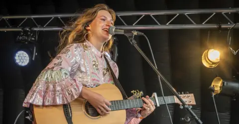 Erica Jean A woman with brown hair is holding a guitar and performing near a microphone.