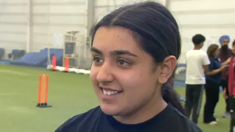 Millie can only be seen from the shoulders up, but she is smiling with her teeth and looking off to the side. She has long black hair which has been tied up into a low ponytail with a middle part. She is wearing a black crew neck sports T-shirt. Out of focus behind her are other players standing together and plastic orange cricket stumps on a green astro floor.