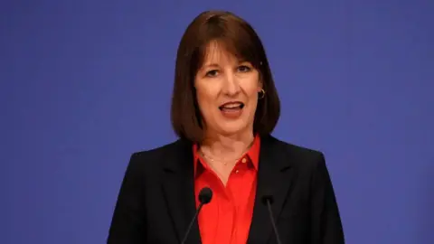 Getty Images Rachel Reeves wearing a red shirt and black blazer. She is talking with two small mics in front of her. The background is blue.