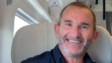 Phil Waddington A man with short brown hair and grey stubble is smiling. He is wearing a dark blue shirt.