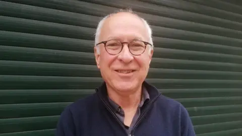 Paul Joslin/BBC Roger King standing against a metal door or wall. He has short cropped white hair and is wearing horn-rimmed glasses and smiling. He is also wearing a blue zipped jumper over a grey shirt.