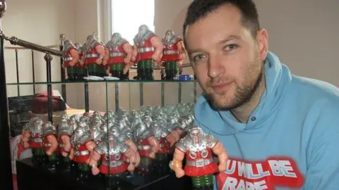 Jamies Moakes wearing a light blue hoody that says "You will be rare" on it. He has short dark hair and a short beard. He is holding a Ram Man action figure, which is muscular and has silver shoulder plates, a red tunic and green trousers. Dozens more Ram Mans are stacked on a glass cabinet behind Moakes.