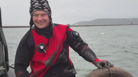 Mallory Haas sitting on the edge of a boat holding a tyre which has been retrieved from the seabed in Plymouth Sound. She is wearing a black and red wetsuit and a black woolly hat with a white skull pattern. She is smiling widely. The sea and the sky are both quite grey.