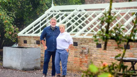 Barbara Leatham Jules and Mark standing in front of the Victorian greenhouse which they are restoring. Jules is wearing blue trousers and a white shirt with her hand in her pocket and her arm around Mark, who is standing to her left wearing blue jeans and a denim shirt. Behind them is a low brick greenhouse with new white roof rafters, but no glass panels fitted yet. The couple are looking at the camera and smiling.