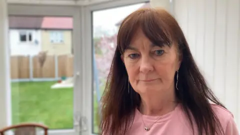 Chris Adamson, a woman with long brown hair, standing in a conservatory with a serious look on her face. She is wearing a pink top and necklace. A lawn, wooden fence and neighbouring houses can be seen through glass doors.