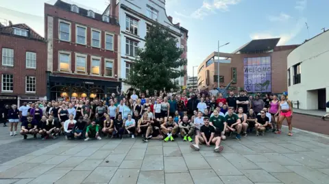 Bristol Run Club A huge group of people are all smiling at the camera. There are a few hundred, they are all in running gear. Some are standing up, some are sitting down. They are on a street with large buildings behind them.