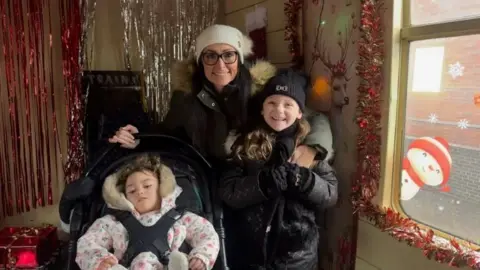 A woman in a black coat and white hat with a girl in a black coat and black hat and a girl in a wheelchair in a pink coat