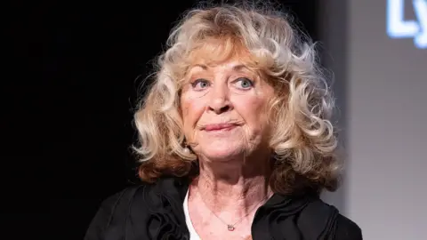 La Plante pictured at an event in 2021, looking away from the camera with a pensive expression on her face, wearing a black top and big blonde curls.