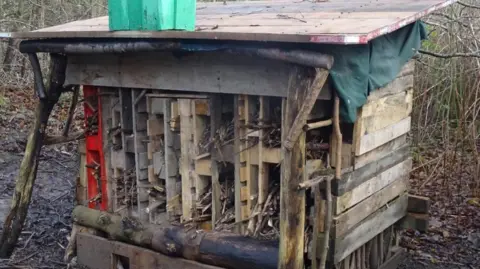Tracy Ostle Set in woodland, the structure is made of wood panels and sticks and features a roof to give insects and other wildlife shelter.