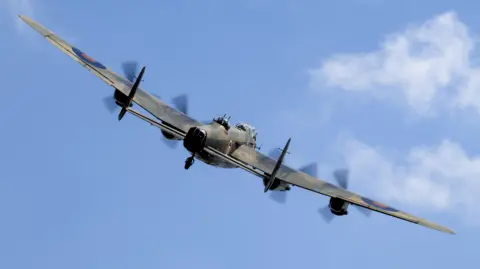 A historic Lancaster bomber plane flies on an angle with four propellers spinning as it takes to the blue skies after a maintenance break.