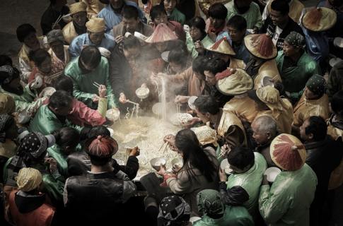 People stand around a giant cauldron eating noodles