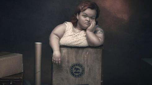 A woman with dwarfism has a bored expression on her face as she stands in a box marked fragile