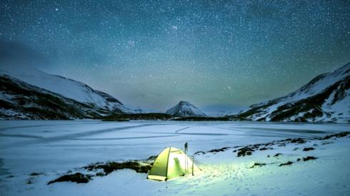 Tent brightly lit from inside sat on the snow below a star covered sky at night