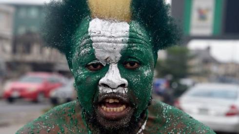 A man with his face painted in the colours of the Nigerian flag – green, white and green – staring into the camera. He is shirtless and outside.