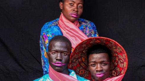 Three models posing wearing colour silk clothing. The male models are wearing pink lipstick.
