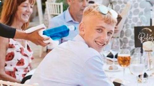 Jake Davies. He has blonde hair and is sat at a table, turning round to smile for the camera. He has sunglasses on his head and is wearing a light blue shirt and beige shorts. 