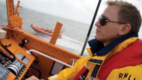 Daniel Craig, wearing sunglasses and a yellow anorak, sits behind the controls on the deck of an orange lifeboat