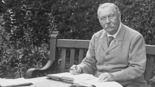 A black and white image of Sir Arthur Conan Doyle sitting outdoors on a bench with an open set of notebooks and a pen in his hand