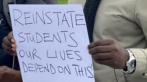 Close up of a man's hand holding a sign saying 'reinstate students our lives depend on this'