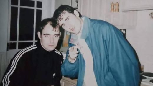 Bonehead and another man at the party. Bonehead is wearing a black Adidas zip-up top, the man wears a blue jacket and points at the camera.