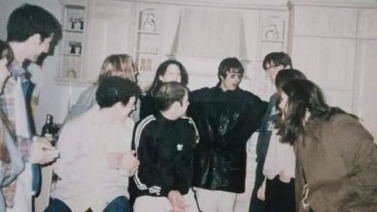 Liam Gallagher wearing glasses and a large black jacket, surrounded by young people in a kitchen, with Bonehead. Everyone is looking at Liam