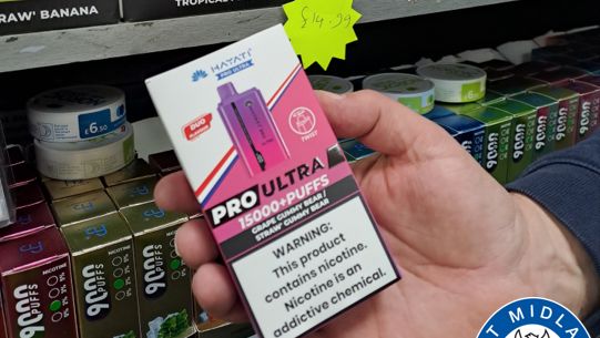 A vape in someone's hand, with others on a shelf behind. The pack says the product offers more than 15,000 puffs and has a warning which says 'this product contains nicotine. Nicotine is an addictive chemical.'