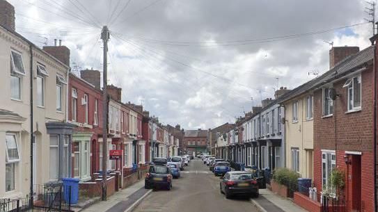 A row of terraced houses with cars parked on either side of the street.