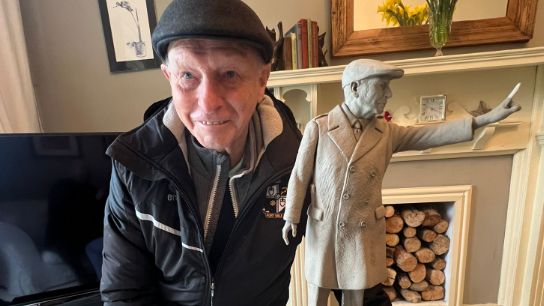 John Rudge, in a flat cap and Port Vale jacket alongside a small version of the statue of himself 