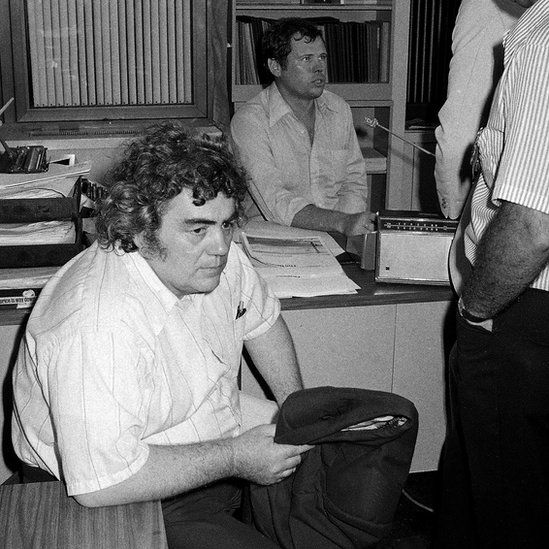 New York Daily News columnist Jimmy Breslin, left, sits at police headquarters in New York after the arrest of David Berkowitz, the Son of Sam serial killer
