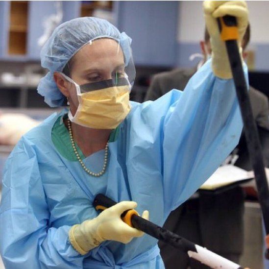Photo of Dr Judy Melinek doing an autopsy wearing protective garments and a string of pearls