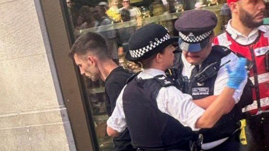 A white man with short dark hair and a black top and trousers stands with his hands behind his back as he is arrested by two officer, with a security guard in a red uniform in the background