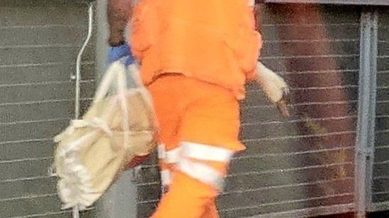 A swan being removed in a bag by a man wearing an orange vest and white hardhat.