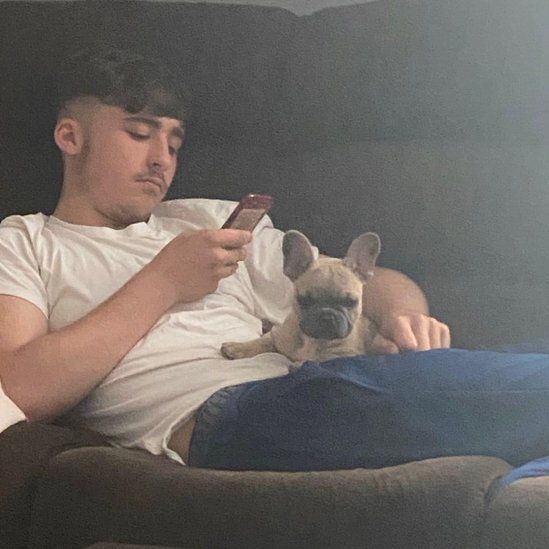 Owen Dunn pictured on a sofa with dog on his lap, looking at his mobile phone