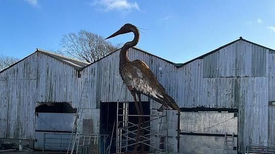 Giant heron sculpture in front of a corrugated iron farm building