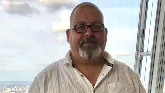 Mark Randle looking at the camera. He is in front of a window showing a view of a town or city and he wears a white shirt and has a short goatee beard and wears glasses with a shaved head