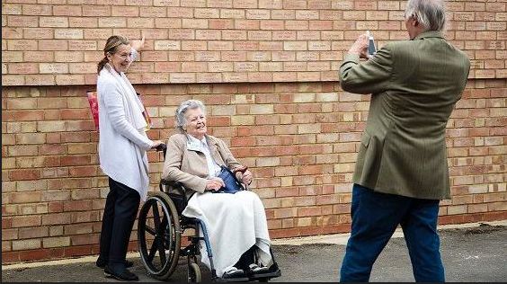 Woman in wheelchair and carer having their photo taken in front of the memorial wall at Bletchley Park