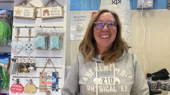 Julie Cooper smiles into the camera in her shop. She has glasses and brown hair and is wearing a grey hoodie. Behind her are a number of items she is selling, including keyrings and ornamental signs.