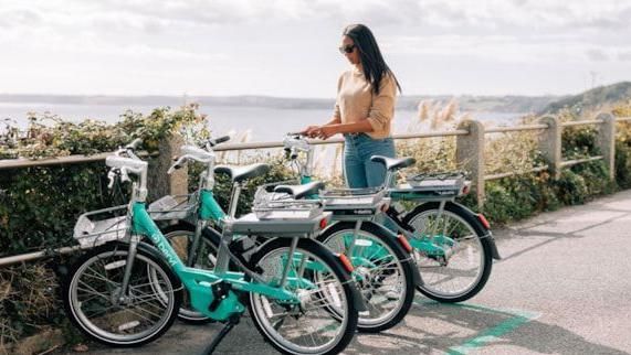 Three light green bikes standing up aside a path overlooking the coast, with a woman with sunglasses stood next to one, trying to get it started 