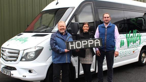 Two men and a woman stand in front of a white bus with the sign 'Hoppa' in their hands.