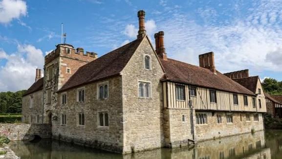 Ightham Mote, a 14th Century stone building in the Tudor style, stands in the centre of a moat on a sunny day with blue skies 