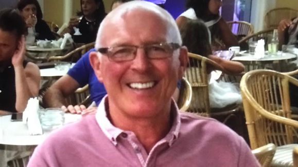 Paddy Murphy, who has short grey hair and glasses and is wearing a pink top, smiles at the camera as he sits in what looks like a venue's dining room