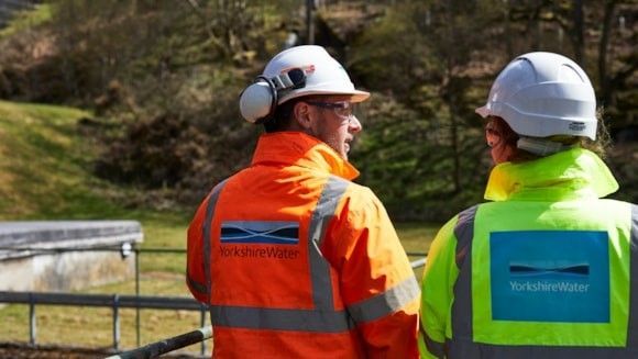 Rear view of two Yorkshire Water employees standing at a railing at a treatment works and looking at each other. Both are wearing brightly-coloured jackets bearing the company's logo and white hard hats.