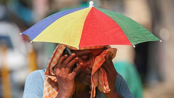 A vendor with an umbrella over his head talks on mobile phone as he wipes his face with a cloth on a hot summer day in Varanasi on May 27, 2024