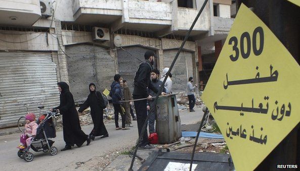 Civilians walk past a sign that reads "300 children without education for two years" in the besieged area of Homs
