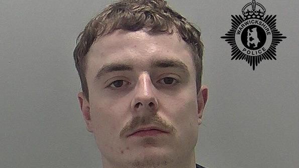 A police mugshot of a man with curly light brown hair and a moustache staring at the camera