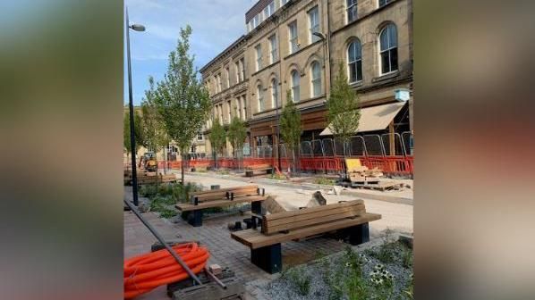 Wooden benches in a street with trees and plants. Orange fencing still surrounds the area